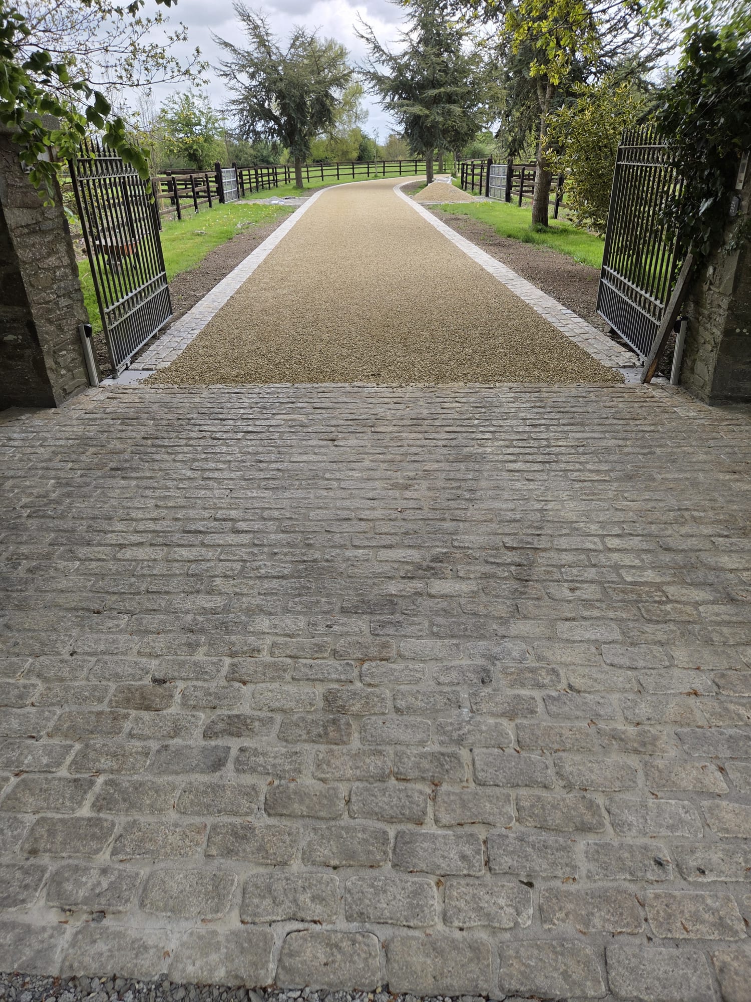 Fresh gravel driveway with a granite sett apron and edged borders at a rural gated entrance with stone pillars and post-and-rail fencing