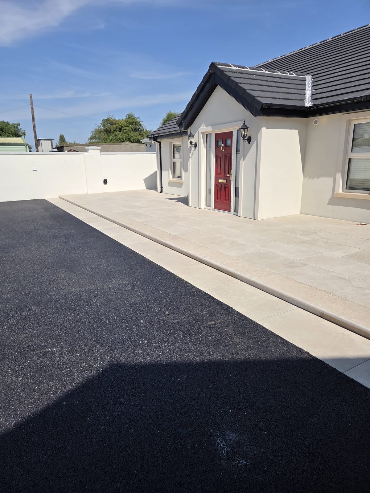 Pale paved apron and step along the front of a white bungalow with a red door, beside the tarmac driveway